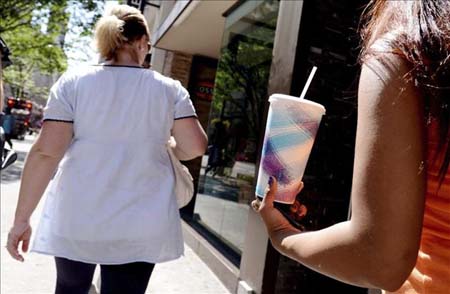 Una mujer con sobrepeso camina por una calle delante de una joven que porta una bebida azucarada.