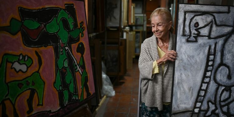 Janine Castes Vigas, the widow of the late Venezuelan artist Oswaldo Vigas (1923-2014) stands next to a painting during an interview with AFP at her husband's studio, in Caracas on January 13, 2023. - "We left it as it was": Oswaldo Vigas' atelier remains intact as it was before his death. A table served as a palette, where the renowned Venezuelan artist mixed the colors, whose work is exhibited in museums and galleries around the world. (Photo by Federico PARRA / AFP)